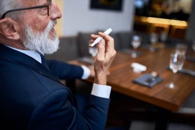 Zengin bir restoranda oturmuş elinde elektronik sigara tutan yaşlı bir adam. Iqos, modern teknolojiler, sigarayı durdurun.