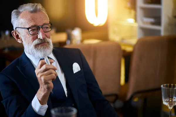 Hombre mayor sosteniendo cigarrillo electrónico sentado en un restaurante rico. IQOS, tecnologías modernas, dejar de fumar — Foto de Stock