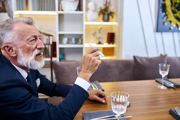 Portrét starší muž šedovlasý sedět v restauraci hledá elektronické cigarety — Stock fotografie