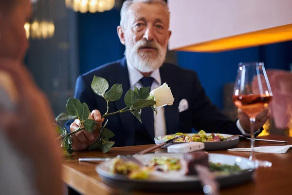 Kärleksfull man som presenterar vit blomma för sin kvinna. Par firar jubileum i restaurang — Stockfoto