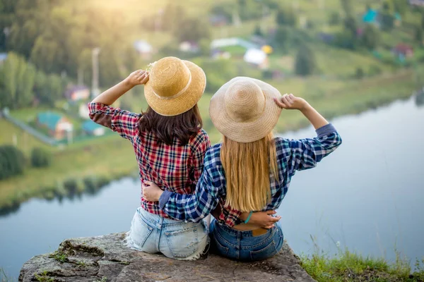 Neşeli kız kardeşler ellerinde şapkalarla dağın tepesinde gün batımının tadını çıkarıyorlar. — Stok fotoğraf
