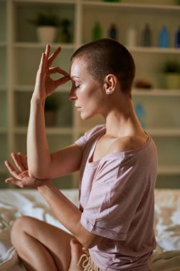 Genç beyaz kadın, parlak yatak odasında meditasyon yapıyor. Yatakta oturuyor.