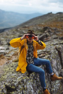 Dürbünle dağda oturan komik adam kameraya bakıyor.