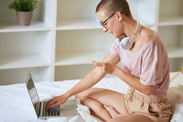 Urocza nowoczesna dziewczyna ze słuchawkami oglądająca w laptopie na łóżku — Zdjęcie stockowe