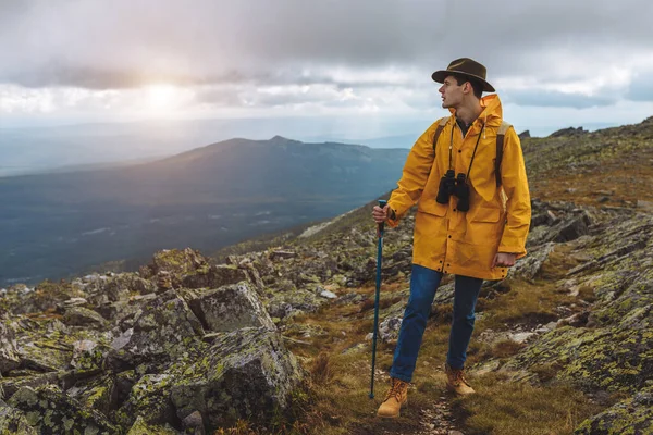 Joven excursionista con sombrero, abrigo amarillo, binocular de pie sobre la roca mirando a un lado — Foto de Stock