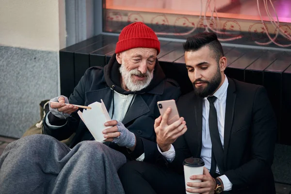 Young kind-hearted businessman and senior beggar look at phone