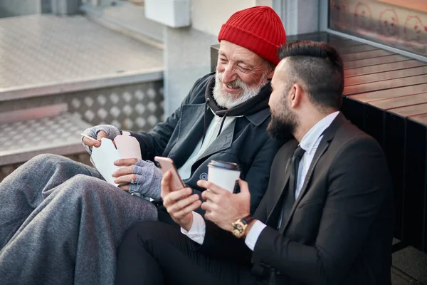Молодой позитивный бизнесмен и зрелый нищий смотрит в телефон — стоковое фото