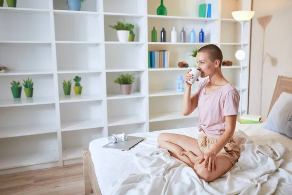 Happy casual pretty caucasian woman with tea sitting on the bed in light room — Stock Photo, Image