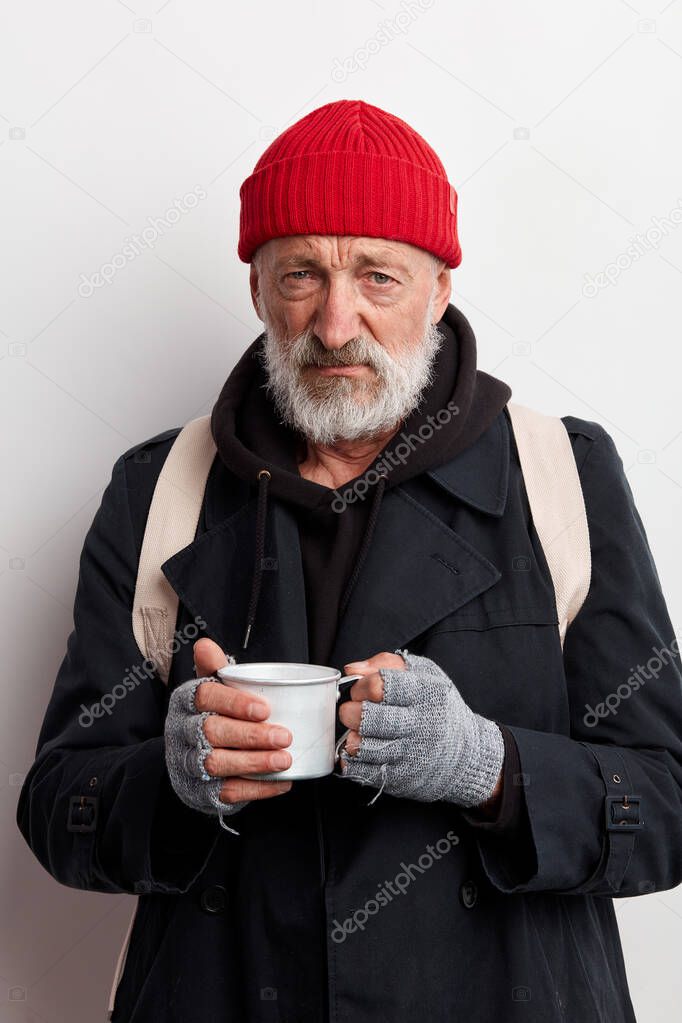 Old bearded beggar covering up in black wear holding mug of hot tea to warm himself in cold day