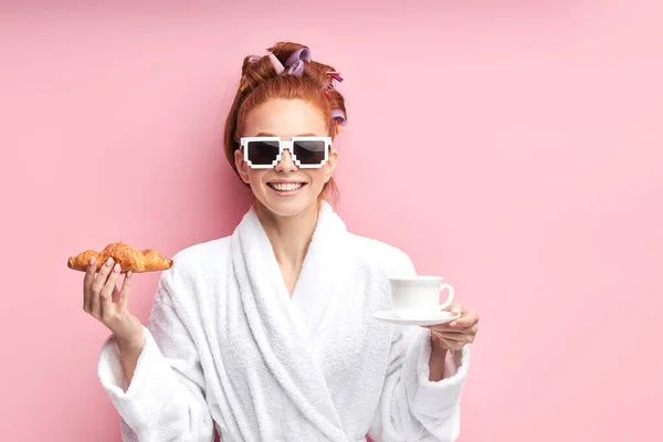 Linda chica pelirroja juguetona en gafas de sol, con albornoz y beber té con croissant — Foto de Stock