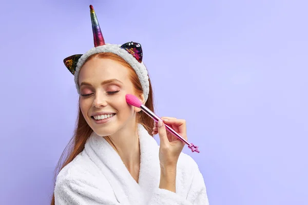 Retrato de mulher atraente aplicando cosméticos com escova isolada no fundo roxo — Fotografia de Stock