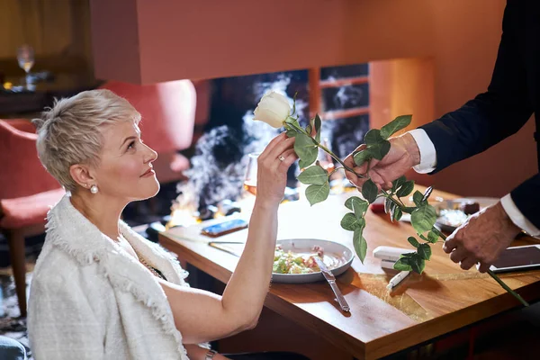 예복을 입은우아 한 남자 가 레스토랑에서 아름다운 금발의 여인을 미소짓게 하는 아름다운 하얀 장미를 주고 있다 — 스톡 사진