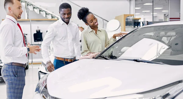 Friendly salesman and beautiful couple in car showroom — Stock Photo, Image