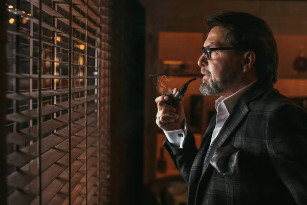 Отражающий хорошо одетый взрослый бородатый мужчина в очках, курящий табачную трубку — стоковое фото
