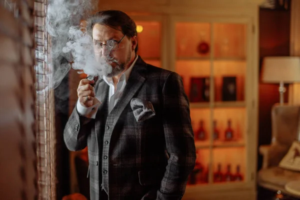 Reflexivo hombre barbudo maduro bien vestido en gafas fumar pipa de tabaco —  Fotos de Stock