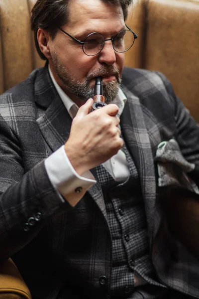Reflexivo hombre barbudo maduro bien vestido en gafas fumar pipa de tabaco — Foto de Stock