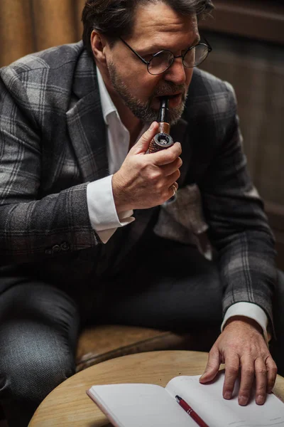 Maduro hombre de negocios fumando pipa de tabaco y tomando notas, trabajando en su oficina . — Foto de Stock