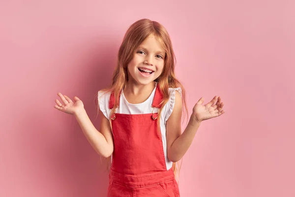 Speels klein meisje in rode overall geïsoleerd op roze achtergrond — Stockfoto