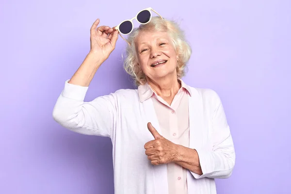 Usměvavá stará dáma drží sluneční brýle a ukazuje palec nahoru — Stock fotografie