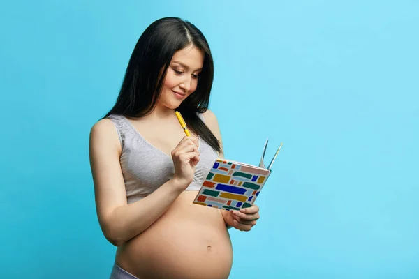 Femeie gravidă în lenjerie de corp scriind în caiet pe perete albastru — Fotografie, imagine de stoc