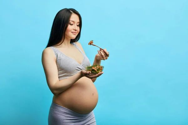 Вагітна струнка жінка з великим животом їсть овочевий салат ізольовано на синій стіні — стокове фото