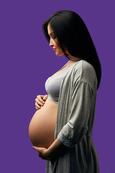 Femeie gravidă care se uită la burtă în timp ce pozează în profil în studioul violet — Fotografie, imagine de stoc