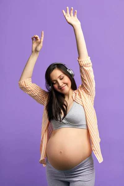 Zâmbind femeie însărcinată în căști dansând în studio pe fundal violet . — Fotografie, imagine de stoc