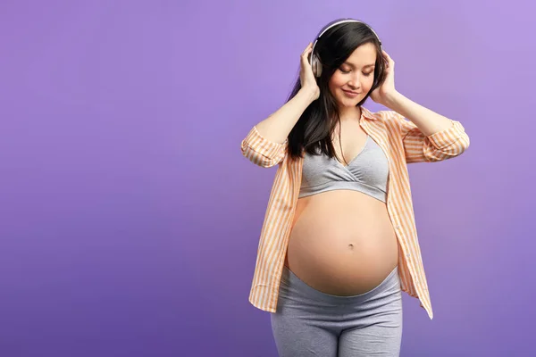 Mujer embarazada sintiéndose saludable escucha música en los auriculares, sobre púrpura — Foto de Stock