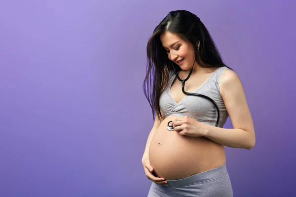 Mujer embarazada en casa llevar escuchando su barriga usando un estetoscopio en el interior — Foto de Stock