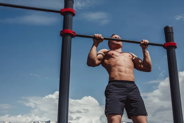 Sports man doing pull-up exercise on a horizontal bar against a blue sky. — Stock Photo, Image