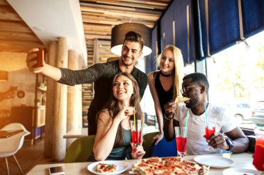 Bir grup arkadaş akıllı bir telefondan selfie çekiyor. Çatıdaki partide pizza yiyen gençler selfie çekiyorlar..
