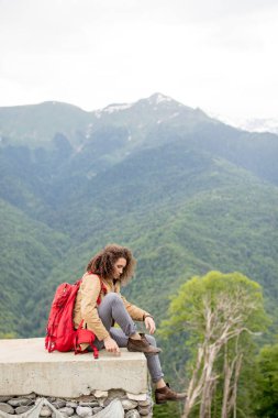 Kırmızı sırt çantalı gezgin dışarıda dinleniyor ve arka planda kayalık dağlar var.