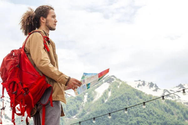 Man Traveler s mapou a červeným batohem vyhledávání umístění venkovní s skalnatými horami na pozadí — Stock fotografie