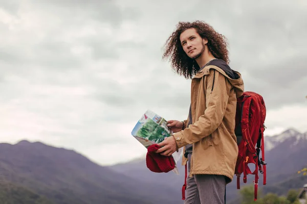 Man Traveler s mapou a červeným batohem vyhledávání umístění venkovní s skalnatými horami na pozadí — Stock fotografie