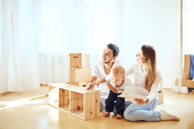 Aile, yeni apartmanın oturma odasında mobilya montajı, arka planda taşınan kutular.