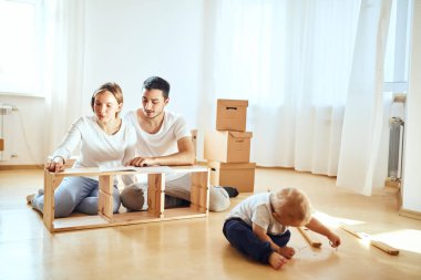 Aile, yeni apartmanın oturma odasında mobilya montajı, arka planda taşınan kutular.