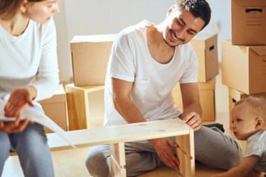 Aile, yeni apartmanın oturma odasında mobilya montajı, arka planda taşınan kutular.
