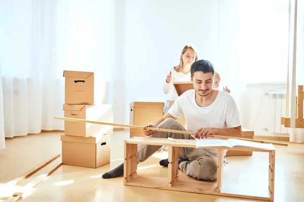 Pai montagem de móveis na sala de estar do novo apartamento, mãe com filho e pilha de caixas móveis no fundo — Fotografia de Stock