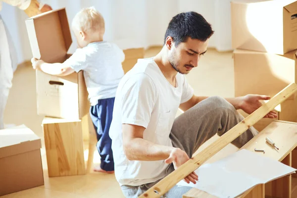 Pai montagem de móveis na sala de estar do novo apartamento, mãe com filho e pilha de caixas móveis no fundo — Fotografia de Stock