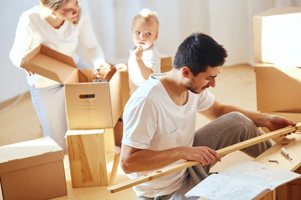 Pai montagem de móveis na sala de estar do novo apartamento, mãe com filho e pilha de caixas móveis no fundo — Fotografia de Stock