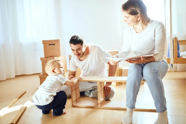 Rodinná čtení instrukce a sestavit nábytek společně v obývacím pokoji nového bytu hromada stěhování boxy na pozadí — Stock fotografie