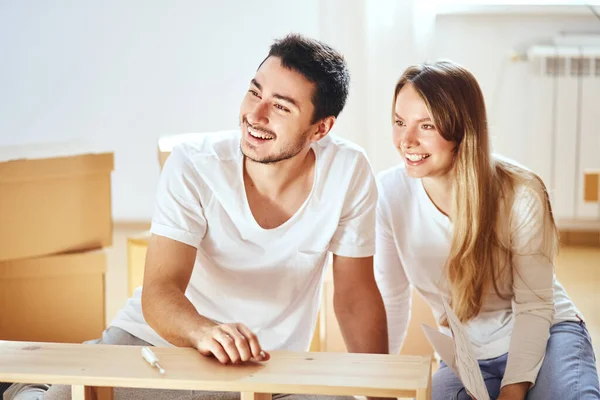 Pareja que ensambla muebles en un nuevo hogar, moviendo cajas en el fondo — Foto de Stock