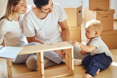 Aile, yeni apartmanın oturma odasında mobilya montajı, arka planda taşınan kutular.