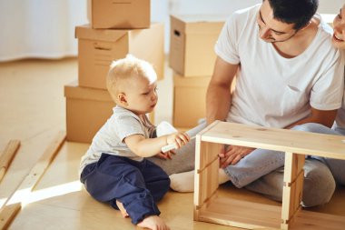Aile, yeni apartmanın oturma odasında mobilya montajı, arka planda taşınan kutular.
