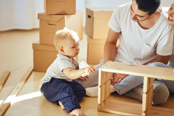 Familia juntos en la sala de estar de nuevo apartamento de montaje de muebles, pila de cajas móviles en el fondo —  Fotos de Stock