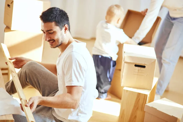 Padre armando muebles en salón de nuevo apartamento, madre con hijo y pila de cajas móviles sobre fondo — Foto de Stock