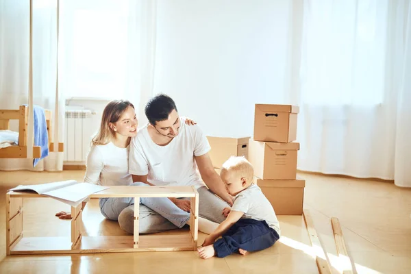 Família juntos na sala de estar do novo apartamento montagem de móveis, pilha de caixas móveis no fundo — Fotografia de Stock