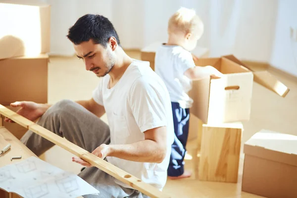 Pai montagem de móveis na sala de estar do novo apartamento, mãe com filho e pilha de caixas móveis no fundo — Fotografia de Stock