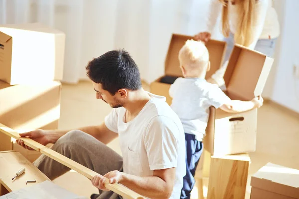 Vader assemblage meubels in de woonkamer van nieuw appartement, moeder met zoon en stapel van verhuisdozen op de achtergrond — Stockfoto