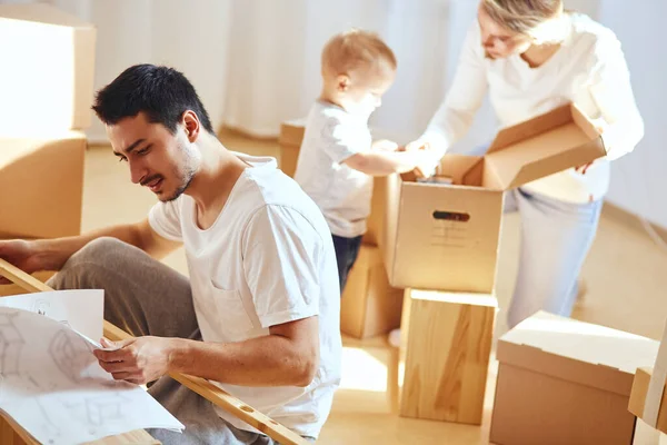 Padre armando muebles en salón de nuevo apartamento, madre con hijo y pila de cajas móviles sobre fondo —  Fotos de Stock